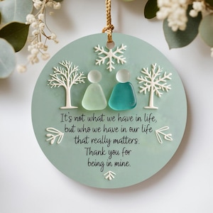 May include: Round, light green ornament with a gold string for hanging. It features two figures made of sea glass, white trees, and a snowflake. The text reads: "It's not what we have in life, but who we have in our life that really matters. Thank you for being in mine."