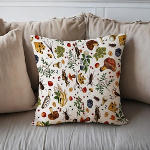 Op de afbeelding: Vierkantig kussen met een crèmekleurige achtergrond met een botanisch patroon van paddenstoelen, insecten, bessen en bladeren in tinten bruin, rood, groen en geel. Het kussen ligt op een bed met neutrale kussens.