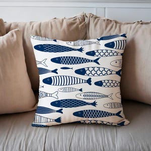 Op de afbeelding: Een vierkant decoratief kussen met een crèmekleurige achtergrond en een patroon van marineblauwe vissen. De vissen variëren in ontwerp, sommige met schubben, andere met strepen en sommige met effen vormen. Het kussen ligt op een neutrale ondergrond.