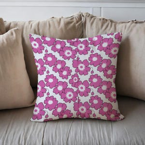 Puede incluir: Cojín cuadrado con un patrón repetido de flores rosas sobre un fondo blanco. Cada flor tiene un centro blanco y un contorno rosa. El cojín está colocado sobre una cama con otros cojines.