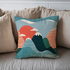May include: Square decorative pillow with a mountain landscape design. The pillow features a vibrant orange sun, blue mountains, and white clouds. The pillow is placed on a light gray surface.