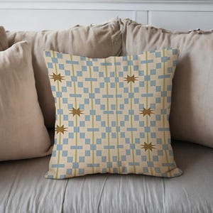 May include: Square cushion with a geometric pattern. The design features a cream background with blue squares, olive green vertical lines, and brown starburst shapes. The cushion is placed on a light grey surface.
