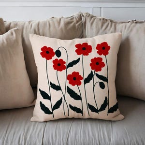 Op de afbeelding: Crèmekleurig vierkant sierkussen met een bloemenontwerp. Het ontwerp bevat rode klaprozen met zwarte stelen en bladeren. Het kussen is omgeven door andere kussens in neutrale tinten.