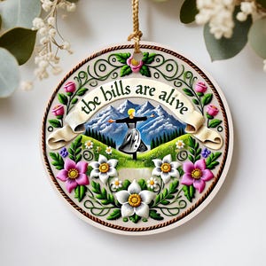 May include: Round wooden ornament with a cream-colored background, featuring a woman with outstretched arms in front of mountains. The text "the bills are alive" is on a banner. The ornament is decorated with pink and white flowers and green leaves.