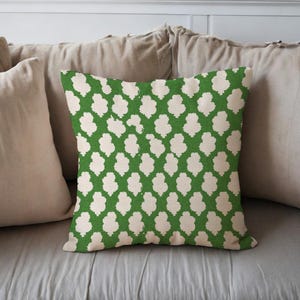 May include: Square decorative pillow with a green background and a repeating pattern of off-white, abstract floral shapes. The pillow is placed on a bed with neutral-colored pillows.
