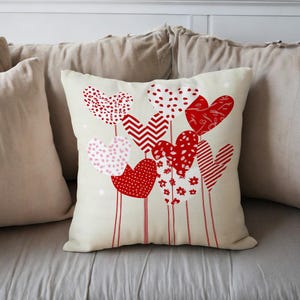 Puede incluir: Un cojín decorativo con un fondo color crema presenta un diseño de corazones rojos y rosas. Los corazones tienen varios patrones, incluyendo lunares, chevrones y diseños florales. El cojín está colocado sobre una superficie gris.