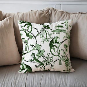 May include: A decorative square pillow with a white background and a green botanical print. The print features monkeys, leopards, flowers, and parasols. The pillow is placed on a light gray surface.
