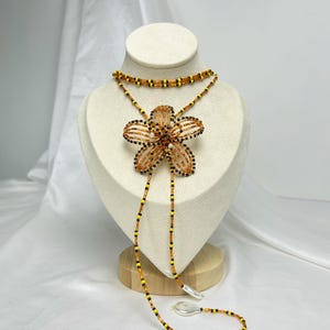 May include: A beaded necklace with a large flower pendant. The flower is orange and black, with a beaded design. The necklace has a choker style with a long beaded strand. The beads are orange, yellow, and black. The necklace is displayed on a white mannequin.