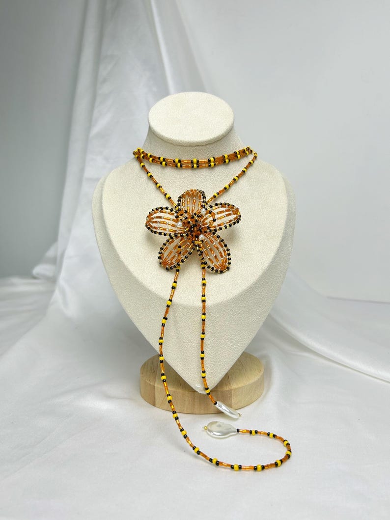 May include: A beaded necklace with a large flower pendant. The flower is orange and black, with a beaded design. The necklace has a choker style with a long beaded strand. The beads are orange, yellow, and black. The necklace is displayed on a white mannequin.