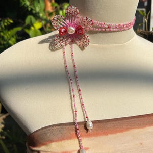 Puede incluir: Un collar tipo gargantilla de cuentas con un motivo de flor de cinco p&eacute;talos hecho de cuentas de semillas rosas, rojas y plateadas. El dise&ntilde;o incluye una perla blanca central, dos hilos de cuentas colgantes que terminan en perlas blancas.