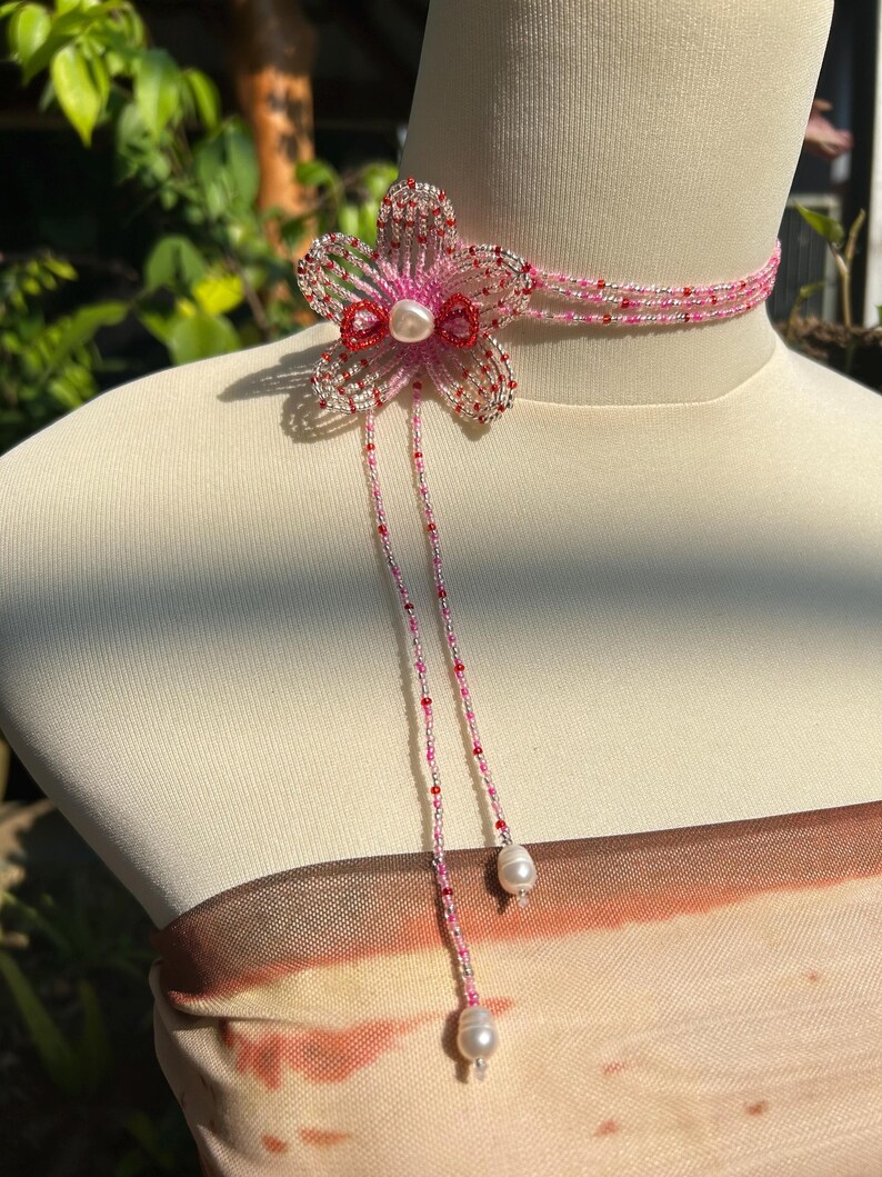 Puede incluir: Un collar tipo gargantilla de cuentas con un motivo de flor de cinco p&eacute;talos hecho de cuentas de semillas rosas, rojas y plateadas. El dise&ntilde;o incluye una perla blanca central, dos hilos de cuentas colgantes que terminan en perlas blancas.