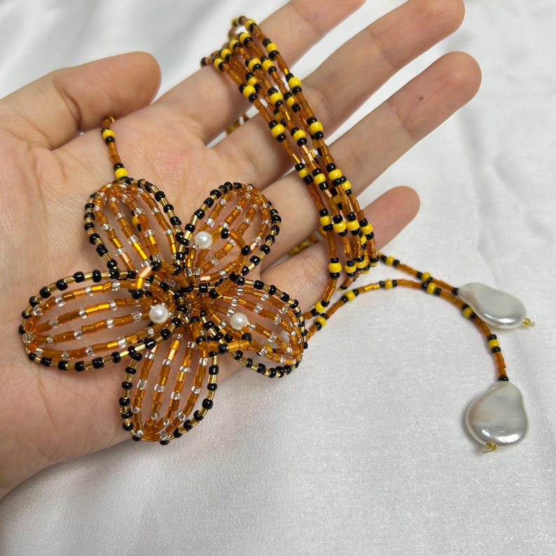May include: A handmade beaded necklace featuring a large flower pendant. The flower is made of orange, black, and clear beads, with small white pearls at the center. The necklace has a multi-strand design with yellow and black beads, and ends with two large, flat, white pearls.