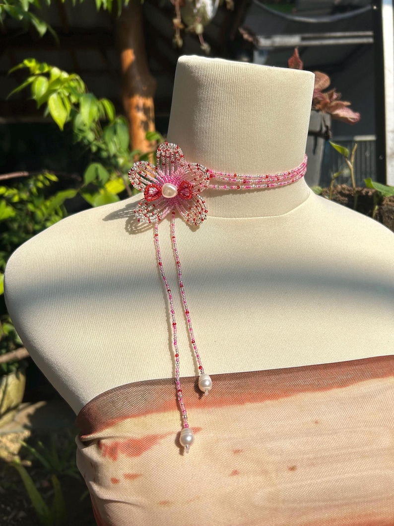 Puede incluir: Un collar tipo gargantilla de varias hileras de cuentas con un motivo floral central de cinco p&eacute;talos hecho de cuentas de vidrio rosas, rojas y transparentes. El dise&ntilde;o incluye una perla blanca en el centro y dos largos colgantes de cuentas que terminan en perlas.