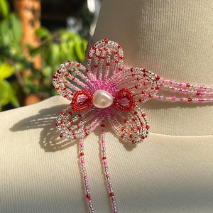 Puede incluir: Un collar tipo gargantilla de cuentas con una flor de cinco p&eacute;talos en el centro, hecha de cuentas de semillas transl&uacute;cidas en rosa, plata y rojo. La flor tiene una cuenta central nacarada y dos largos hilos de cuentas que cuelgan hacia abajo.