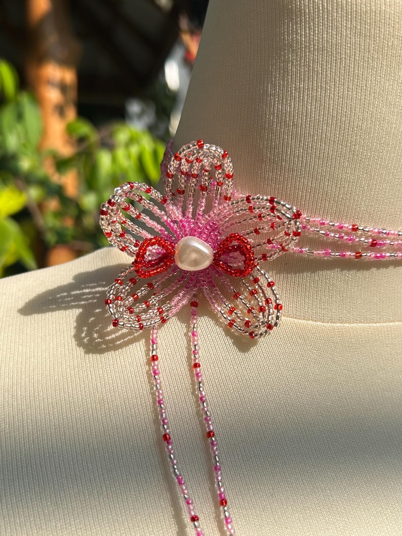 Puede incluir: Un collar tipo gargantilla de cuentas con una flor de cinco p&eacute;talos en el centro, hecha de cuentas de semillas transl&uacute;cidas en rosa, plata y rojo. La flor tiene una cuenta central nacarada y dos largos hilos de cuentas que cuelgan hacia abajo.