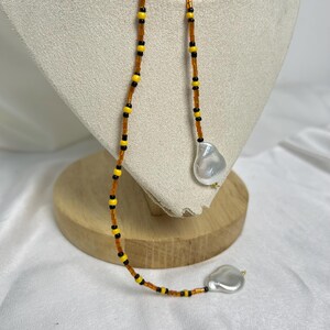 May include: A beaded necklace with a unique design. The necklace features a strand of small, orange beads interspersed with yellow and black beads. Two large, irregular-shaped, white pearls dangle from the ends of the necklace. The necklace is displayed on a cream-colored stand.