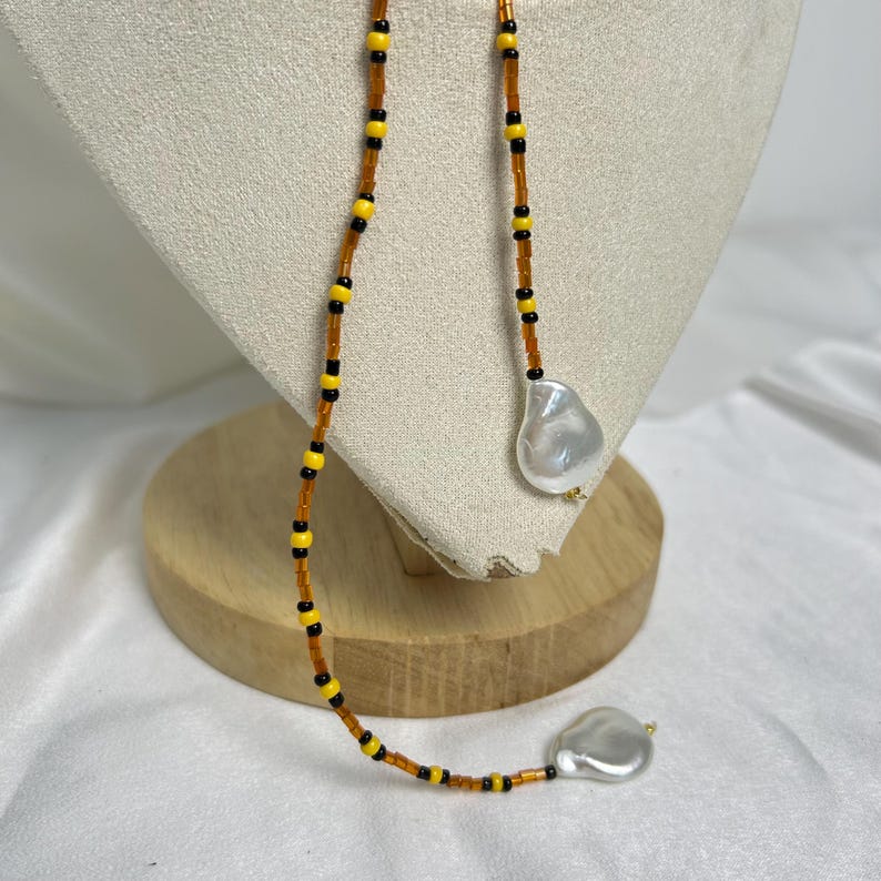 May include: A beaded necklace with a unique design. The necklace features a strand of small, orange beads interspersed with yellow and black beads. Two large, irregular-shaped, white pearls dangle from the ends of the necklace. The necklace is displayed on a cream-colored stand.