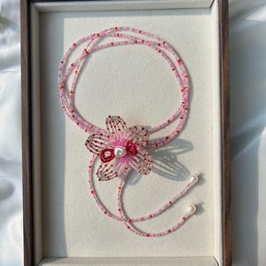 Puede incluir: Un collar de cuentas de varias hebras con un colgante central en forma de flor hecho de cuentas rosas, rojas y plateadas con una perla blanca en el centro. El collar incluye dos extremos colgantes terminados con perlas blancas.