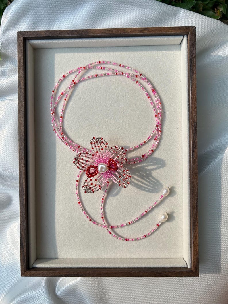 Puede incluir: Un collar de cuentas de varias hebras con un colgante central en forma de flor hecho de cuentas rosas, rojas y plateadas con una perla blanca en el centro. El collar incluye dos extremos colgantes terminados con perlas blancas.