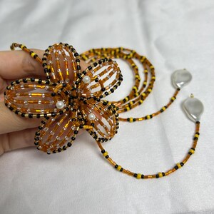 May include: A handmade beaded flower pendant necklace. The flower is made of orange, black, and gold beads with small white pearl accents. The necklace has a long strand of beads in orange, yellow, and black, with two large, irregular white pearl drops.