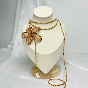 May include: A beaded necklace with a large flower pendant. The flower is made of orange and black beads. The necklace has a long, flowing design with a pear-shaped bead at the end. The necklace is displayed on a mannequin.