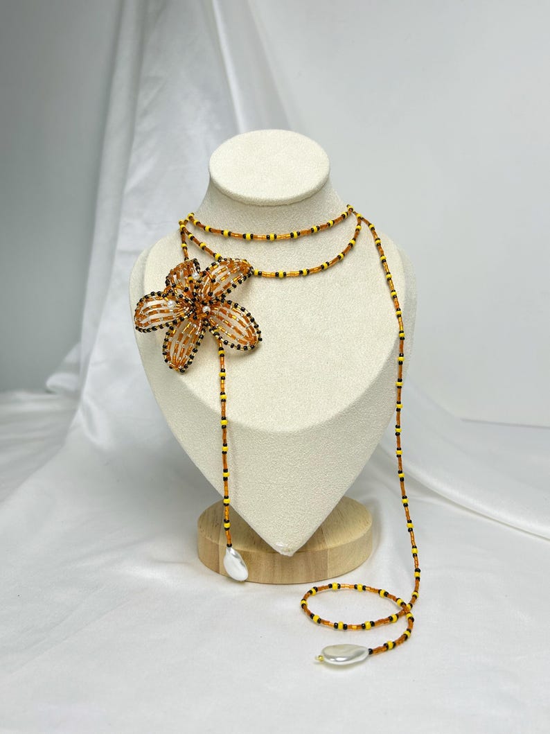 May include: A beaded necklace with a large flower pendant. The flower is made of orange and black beads. The necklace has a long, flowing design with a pear-shaped bead at the end. The necklace is displayed on a mannequin.