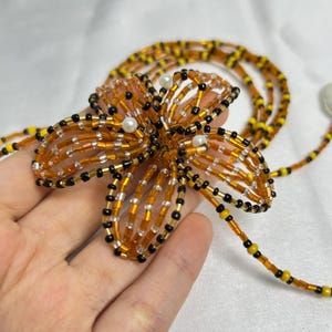 May include: A beaded flower necklace with a detailed design. The flower is made of orange, black, and clear beads, with small white pearls at the center. The necklace chain features yellow and black beads.