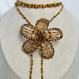 May include: A beaded necklace featuring a large flower pendant. The flower is made of orange, black, and yellow beads, with small white beads in the center. The necklace has a double strand design with matching beadwork.