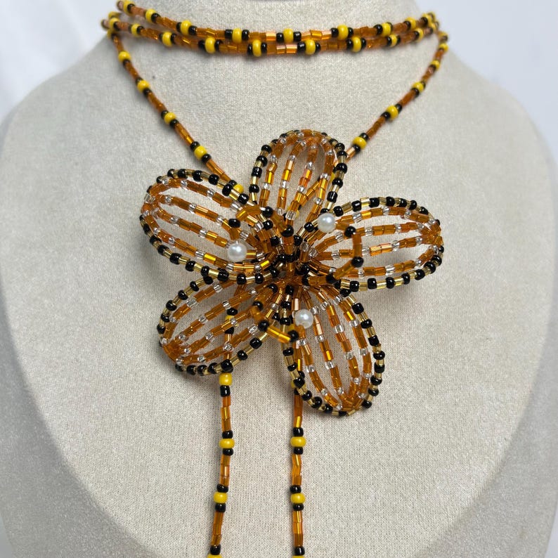 May include: A beaded necklace featuring a large flower pendant. The flower is made of orange, black, and yellow beads, with small white beads in the center. The necklace has a double strand design with matching beadwork.
