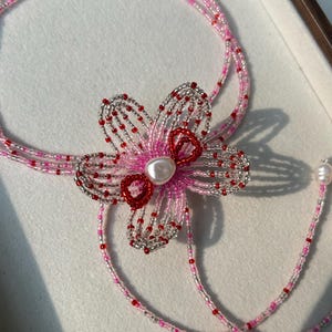 Puede incluir: Un collar de cuentas de varias hebras con un motivo floral central hecho de cuentas de semillas transl&uacute;cidas rosas, transparentes y rojas. El centro de la flor est&aacute; acentuado con una cuenta blanca tipo perla y dos peque&ntilde;os bucles rojos.