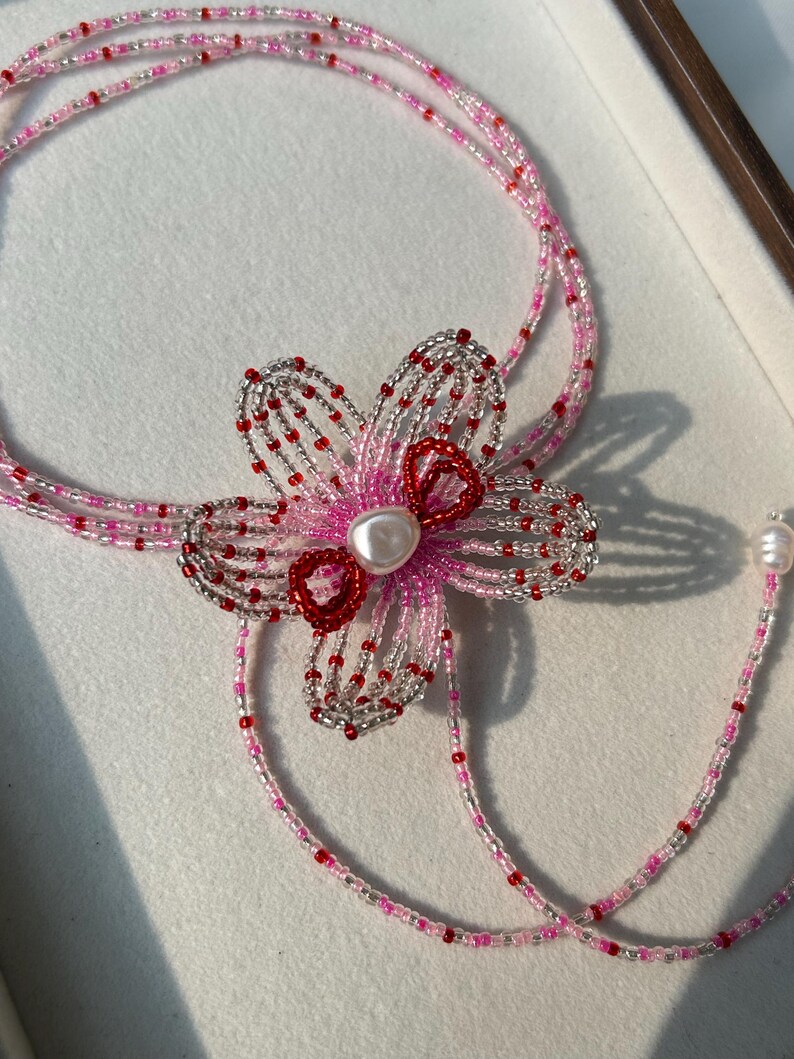 Puede incluir: Un collar de cuentas de varias hebras con un motivo floral central hecho de cuentas de semillas transl&uacute;cidas rosas, transparentes y rojas. El centro de la flor est&aacute; acentuado con una cuenta blanca tipo perla y dos peque&ntilde;os bucles rojos.