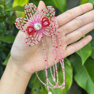 Puede incluir: Un accesorio de flor con cuentas que presenta cinco p&eacute;talos hechos de cuentas de vidrio transparentes, rosas y rojas. El centro de la flor est&aacute; adornado con una cuenta blanca similar a una perla. Largos hilos de cuentas rosas y rojas cuelgan.