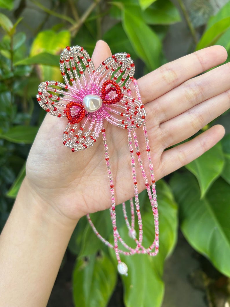 Puede incluir: Un accesorio de flor con cuentas que presenta cinco p&eacute;talos hechos de cuentas de vidrio transparentes, rosas y rojas. El centro de la flor est&aacute; adornado con una cuenta blanca similar a una perla. Largos hilos de cuentas rosas y rojas cuelgan.