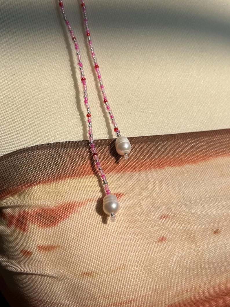 Puede incluir: Un top sin tirantes con un degradado en tonos beige, marr&oacute;n y naranja, acentuado por dos delicados cordones de cuentas. Cada cord&oacute;n est&aacute; compuesto por peque&ntilde;as cuentas rosas, rojas y plateadas, terminando en una perla de agua dulce blanca.