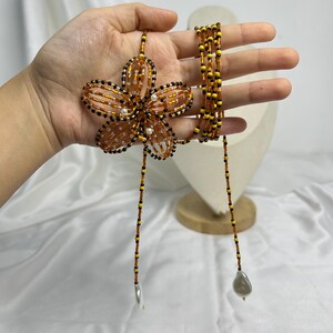 May include: A beaded necklace featuring a large, orange flower pendant with black and white accents. The necklace has a long, beaded strand with a silver-colored charm and a gray pearl-shaped bead at the ends. The flower pendant is approximately 4 inches in diameter.