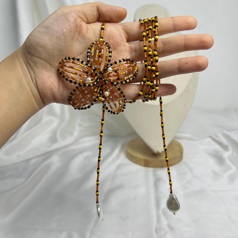 May include: A beaded necklace featuring a large, orange flower pendant with black and white accents. The necklace has a long, beaded strand with a silver-colored charm and a gray pearl-shaped bead at the ends. The flower pendant is approximately 4 inches in diameter.