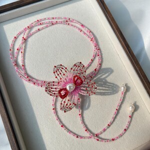 Puede incluir: Un collar de varias hebras de cuentas con un motivo floral central hecho de cuentas de semillas plateadas, rosas y rojas. El centro de la flor est&aacute; acentuado con una perla blanca. El collar termina con dos perlas blancas colgantes.