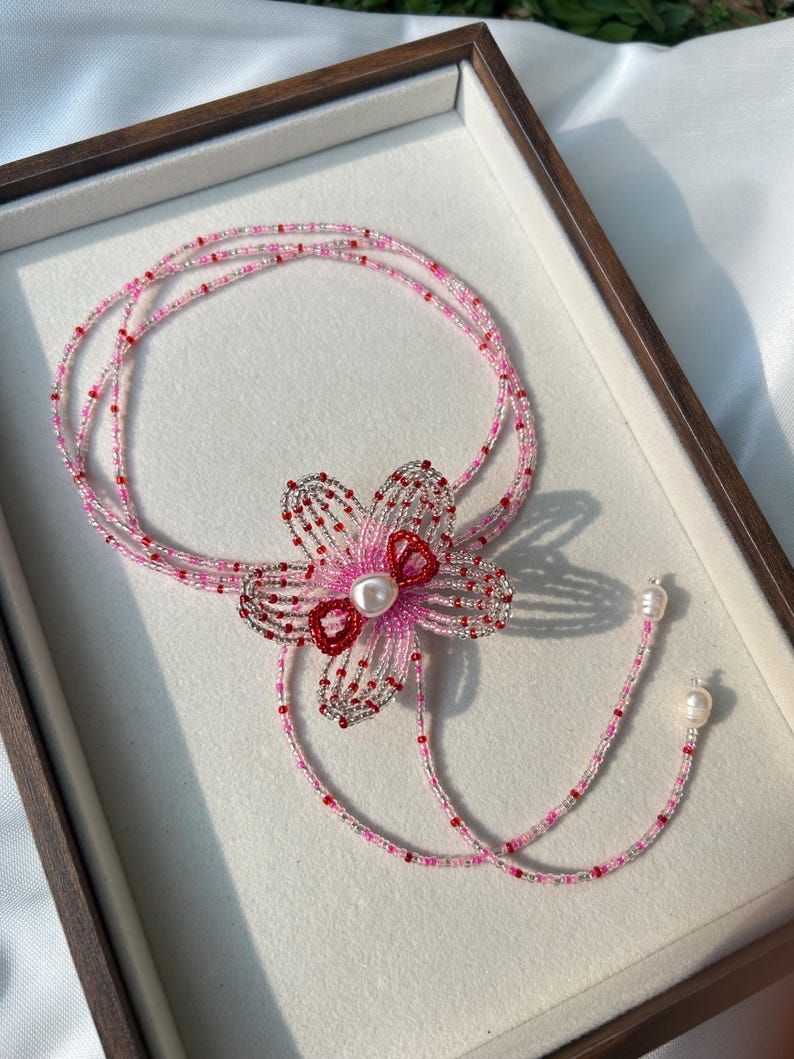 Puede incluir: Un collar de varias hebras de cuentas con un motivo floral central hecho de cuentas de semillas plateadas, rosas y rojas. El centro de la flor est&aacute; acentuado con una perla blanca. El collar termina con dos perlas blancas colgantes.
