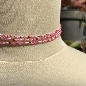 Puede incluir: Un collar tipo gargantilla de m&uacute;ltiples hilos con peque&ntilde;as cuentas uniformes en tonos alternos de rosa transl&uacute;cido, rosa intenso opaco, plata met&aacute;lica y rojo oscuro, dispuestas en un patr&oacute;n repetitivo a lo largo de tres capas circulares.