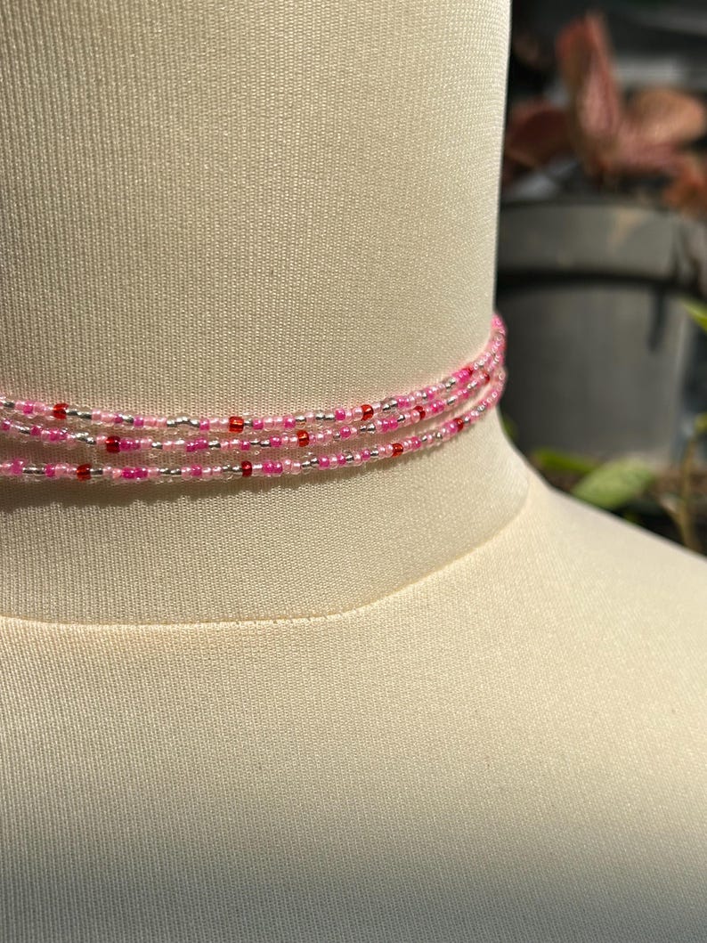 Puede incluir: Un collar tipo gargantilla de m&uacute;ltiples hilos con peque&ntilde;as cuentas uniformes en tonos alternos de rosa transl&uacute;cido, rosa intenso opaco, plata met&aacute;lica y rojo oscuro, dispuestas en un patr&oacute;n repetitivo a lo largo de tres capas circulares.