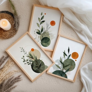 Op de afbeelding: Drie ingelijste botanische kunstprints met aquarelontwerpen. Elke print heeft groene bladeren, oranje cirkels en abstracte vormen. De frames zijn van licht hout. De prints zijn gerangschikt op een neutraal getinte ondergrond.