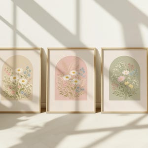 May include: Three framed floral art prints. Each print features a bouquet of flowers within an arched shape. The backgrounds are in soft pastel colours: beige, pink, and green. The frames are a light wood colour, and the overall aesthetic is delicate and botanical.