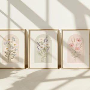 May include: Three framed floral art prints. The prints feature botanical illustrations of daisies, lavender, and cosmos flowers. Each print is set against a neutral background within a gold frame. The artwork is displayed against a white wall with shadows.