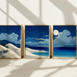 Peut inclure: Trois impressions d'art encadrées représentant un paysage côtier. Chaque impression montre un ciel nocturne avec des nuages, une plage de sable et un plan d'eau. L'œuvre utilise des nuances de bleu, blanc et beige, avec des cadres dorés.