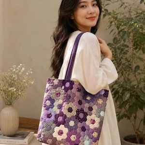 Pode incluir: Uma bolsa tote acolchoada com um design de patchwork de hexágonos de tecido em tons de roxo, lavanda e creme dispostos em padrões florais. A bolsa tem duas alças de ombro de tecido roxo correspondentes e uma textura macia e acolchoada.