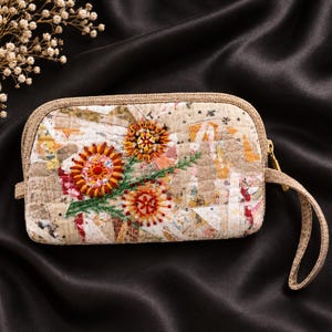 May include: A rectangular quilted zipper pouch featuring a beige and white patchwork fabric base with three embroidered orange and yellow flower motifs, green stems, and a matching beige wrist strap with a gold-toned metal zipper pull.