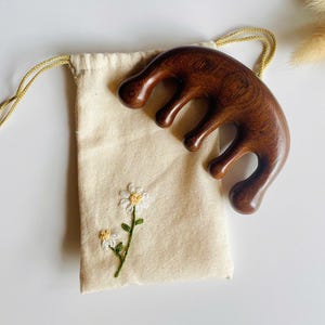 May include: A dark brown, curved wooden massage tool with four rounded prongs, positioned next to a small, off-white fabric drawstring pouch featuring an embroidered daisy flower design with green stems and white petals.