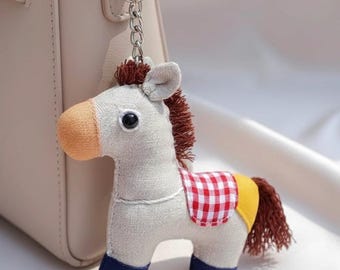 May include: A plush horse keychain featuring a light beige body, a tan muzzle, and dark blue hooves. The horse has a brown yarn mane and tail, and wears a red and white checkered saddle blanket with a yellow accent. It hangs from a silver metal chain.