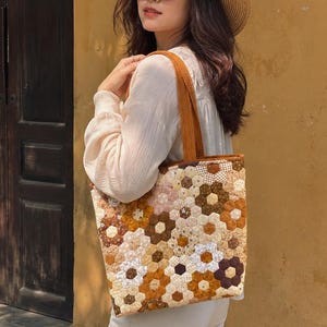 Pode incluir: Uma bolsa tote com um design de patchwork de peças de tecido hexagonais em tons de castanho, bege, creme e branco. A bolsa tem duas alças de ombro em tecido castanho sólido e uma forma retangular, feita de um tecido acolchoado e estampado.