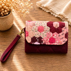 Può includere: Un portafoglio trapuntato bordeaux con cinturino da polso. La patta presenta un design patchwork di forme esagonali rosa e bordeaux con motivi floreali. Il portafoglio ha una chiusura color ottone ed è su una superficie di legno.